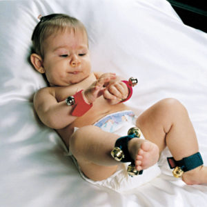 Bebê deitado sob um tapete branco, com Guizos nos pés e nas mãos. Com a mão direita, toca a pulseira que está em seu pulso esquerdo.