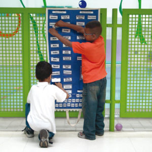 Dois meninos estão de costas, em frente ao painel do Passatempo. O da esquerda, está agachado e toca um dos cartões, enquanto o que está à direita, em pé, está com ambas as mãos em um dos cartões.