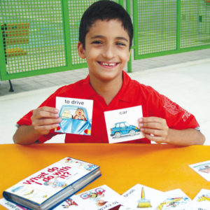 Sentado em frente à uma mesa amarela, um menino de aproximadamente 10 anos, cabelos escuros, curtos e sorriso no rosto, segura um cartão com a mão esquerda e outro com a direita.
