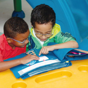 Sentados em frente à uma mesa amarela, dois meninos de aproximadamente 6 anos, cabelos escuros, curtos e óculos, tocam com as mãos, uma página do livro Ajudando a Mamãe.