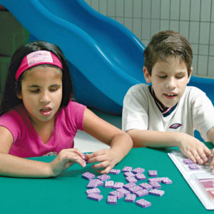 Uma menina e um menino de aproximadamente 7 anos, sentados lado a lado, em frente à uma mesa verde. Ambos têm nas mãos, algumas celas do Brailito.