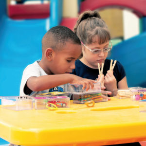 Um menino e uma menina de aproximadamente 5 anos, sentados em frente à uma mesa amarela. O menino está com a mão sobre a caixinha de plástico e ao lado dele, a menina tem nas mãos, dois pregadores de plástico.