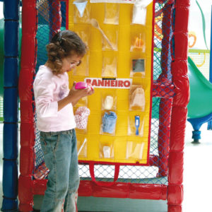 Menina de aproximadamente 6 anos, está em pé, em frente ao tapete amarelo do brinquedo Como Gente Grande. Na mão direita, segura um dos objetos que compõem o brinquedo.