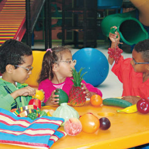 Dois meninos e uma menina de aproximadamente 5 anos, sentados em frente a uma mesa amarela, onde estão as frutas de plástico e a sacolinha do brinquedo Feirinha.