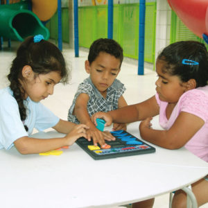 Duas meninas e um menino, de aproximadamente 3 anos, estão sentados, lado a lado, em frente à uma mesa branca. No centro da mesa, o brinquedo Form Color. Os três têm nas mãos, uma forma geométrica.