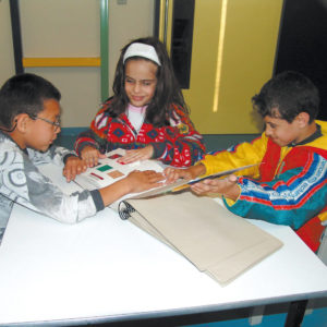 Dois meninos e uma menina sentados em frente à uma mesa branca, tocam páginas do Livro Sensorial.