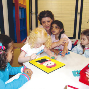 Três meninas de aproximadamente 5 anos, sentadas lado a lado, em frente à uma mesa branca. A que está no meio, cabelos claros e cacheados, tem nas mãos, peças de encaixe do brinquedo Toninho.