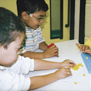 Dois meninos de aproximadamente 5 anos, sentados lado a lado, em frente à uma mesa branca, onde está o Brailindo. O que está à esquerda, toca pinos amarelos espalhados sobre a mesa e ao seu lado, um menino tem nas mãos uma caixinha vermelha.