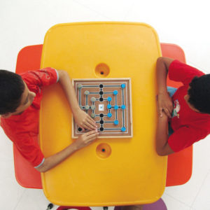 Dois meninos, sentados um em frente ao outro, em frente à uma mesa amarela, onde está o tabuleiro da Trilha. O que está à esquerda, toca uma peça, enquanto o outro, observa.