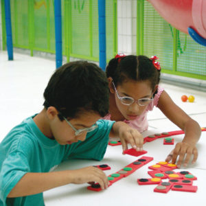 Um menino e uma menina de aproximadamente 5 anos, sentados lado a lado. Tocam peças do Dominó de Texturas.