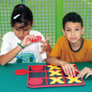 Uma menina e um menino, sentados lado a lado, em frente ao tabuleiro de Jogo da Velha. Ela segura um círculo vermelho e ele, toca o X no tabuleiro.