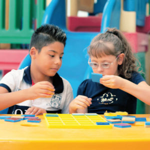 Um menino e uma menina de aproximadamente 6 anos, sentados em frente à uma mesa amarela, onde está o Jogo da Memória. Ela segura um quadrado azul com a mão esquerda, enquanto ele observa.