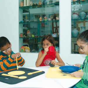 Duas meninas e um menino sentados ao redor de uma mesa branca. Da esquerda para a direita, menino em frente à prancha de alimentação azul; menina com caneca vermelha na mão direita; menina em frente ao prato azul, sob prancha de alimentação amarela.